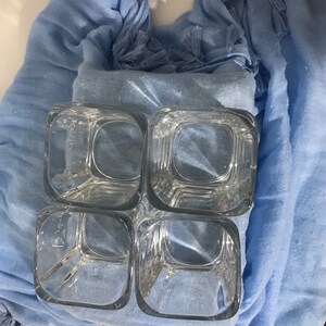 May include: Four clear glass square tumblers with a textured surface, stacked on top of each other, on a light blue fabric background.