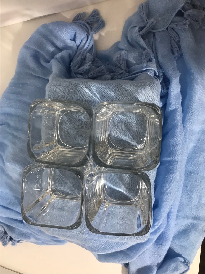 May include: Four clear glass square tumblers with a textured surface, stacked on top of each other, on a light blue fabric background.