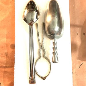 May include: A collection of vintage kitchen utensils, including a long-handled spoon, ice cream scoop, and sugar tongs. The items are made of shiny, reflective metal and are arranged on a white surface. The ice cream scoop has a decorative, twisted handle.