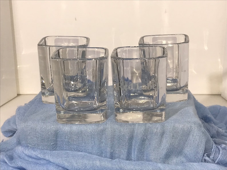 May include: Four clear glass tumblers with a square shape. The tumblers are etched with the words "Johnnie Walker Red Label".