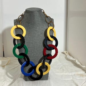 May include: A bold necklace featuring large, interlocking rings in a variety of colors, including yellow, black, green, blue, and red. The rings are connected by a gold-toned chain. The necklace is displayed on a gray mannequin head.
