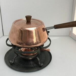 May include: A copper fondue pot with a wooden handle and lid, sitting on a black metal stand with a circular base.