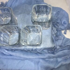 May include: Four clear glass square tumblers with a textured surface, sitting on a blue fabric with tassels.
