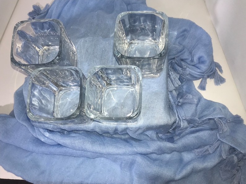 May include: Four clear glass square tumblers with a textured surface, sitting on a blue fabric with tassels.
