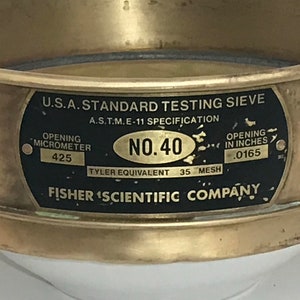 May include: A gold-colored metal sieve with a black label that reads "U.S.A. STANDARD TESTING SIEVE", "A.S.T.M.E-11 SPECIFICATION", "NO.40", "TYLER EQUIVALENT 35 MESH", "FISHER SCIENTIFIC COMPANY", "OPENING MICROMETER 425", and "OPENING IN INCHES .0165".