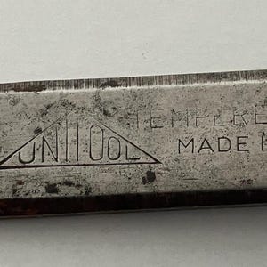 May include: Close-up of a vintage metal tool with the brand name "UNI TOOL" inside a triangle. The word "TEMPERED" is etched above "MADE IN". The tool has a worn, textured appearance.