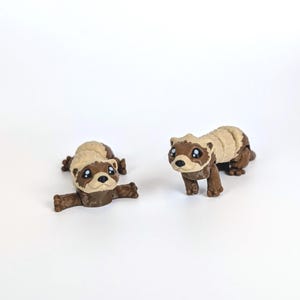 May include: Two small, brown and tan ferret figurines. One is lying down with its arms outstretched, and the other is standing. Both have large, dark eyes and are set against a white background.