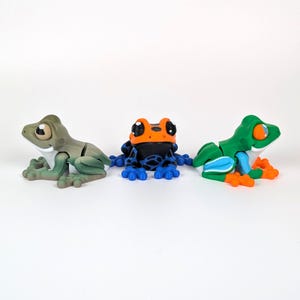 May include: Three colorful frog figurines. One is olive green, another is black with blue spots and orange accents, and the third is green with blue and orange details. The frogs are posed on a white surface.