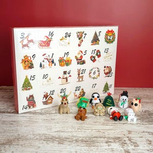 May include: A Christmas advent calendar with numbered doors and festive illustrations. The calendar features miniature figurines of a reindeer, penguin, snowman, and Santa Claus. The background is a red wall and a wooden surface.