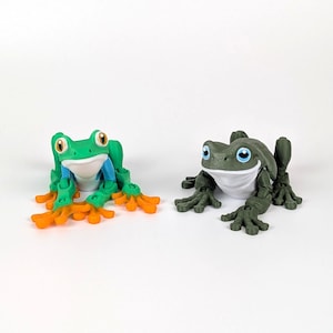 May include: Two toy frog figurines. One is green, blue, and orange, the other is dark green. Both have large eyes and are posed as if sitting. The frogs are on a white background.