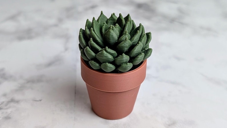 May include: A small, artificial succulent plant in a terracotta-colored pot. The succulent has multiple green leaves arranged in a rosette shape. The pot is cylindrical with a slightly flared rim. The background is a marble pattern.