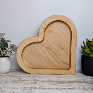 May include: A heart-shaped wooden tray with a raised edge and a natural wood grain finish. The tray is light brown and sits on a light-colored surface. The tray is likely used for serving or display.