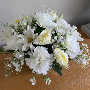 May include: A white flower arrangement featuring a mix of white carnations, daisies, and roses. The arrangement is displayed in a clear vase on a wooden table.
