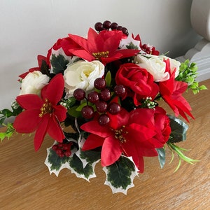 May include: A festive floral arrangement featuring red poinsettias, white roses, and red berries. Green leaves and holly accents complete the holiday-themed display. The arrangement is set on a wooden surface.