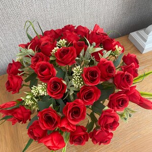 May include: A vibrant arrangement of red roses, accented with green leaves and small white flowers. The bouquet is densely packed, creating a full, lush appearance. The flowers are set on a wooden surface.