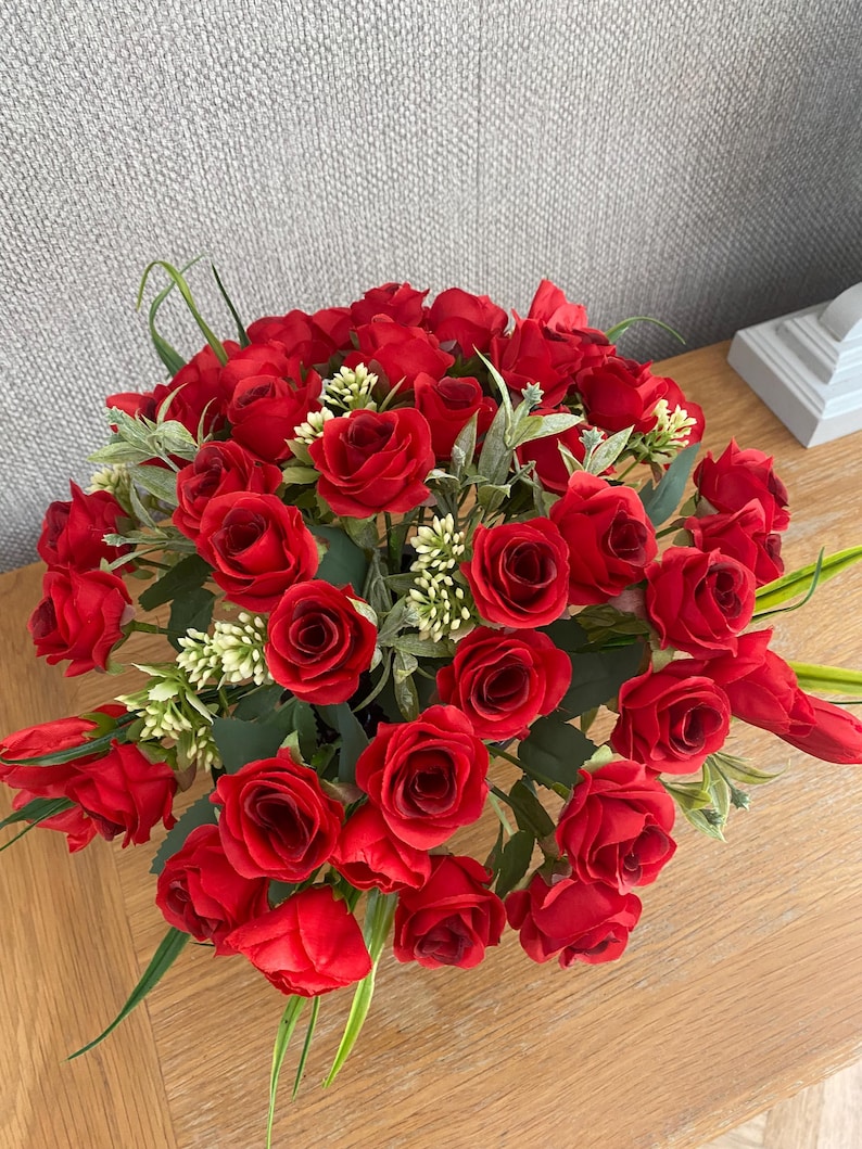 May include: A vibrant arrangement of red roses, accented with green leaves and small white flowers. The bouquet is densely packed, creating a full, lush appearance. The flowers are set on a wooden surface.