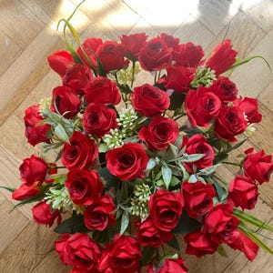 May include: A vibrant arrangement of red roses, with green leaves and small white flowers, creating a full, lush display. The roses are in various stages of bloom, adding depth and texture to the floral arrangement.