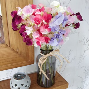 Puede incluir: Un ramo de flores de guisante de olor p&uacute;rpura, rosa y blanco en un jarr&oacute;n de vidrio transparente con una cinta marr&oacute;n alrededor del cuello.
