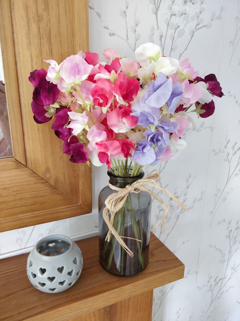 Puede incluir: Un ramo de flores de guisante de olor p&uacute;rpura, rosa y blanco en un jarr&oacute;n de vidrio transparente con una cinta marr&oacute;n alrededor del cuello.