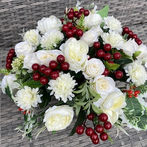 May include: A close-up of a floral arrangement with white roses, chrysanthemums, and red berries. The artificial flowers are densely packed, with the berries adding a splash of colour. Green foliage is also visible within the arrangement.