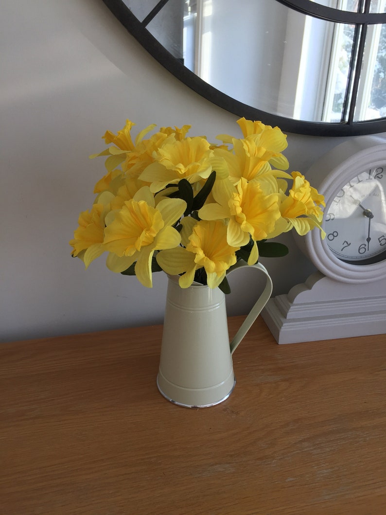 May include: A bouquet of yellow daffodils in a cream-colored ceramic pitcher on a wooden table.