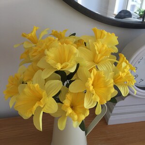 May include: A bouquet of yellow daffodils in a cream-colored ceramic pitcher. The pitcher has a handle and a silver rim.