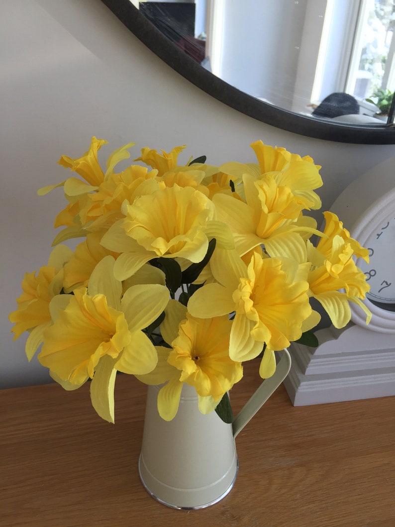 May include: A bouquet of yellow daffodils in a cream-colored ceramic pitcher. The pitcher has a handle and a silver rim.