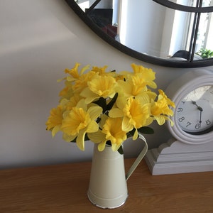 May include: A bouquet of yellow daffodils in a cream-colored metal pitcher on a wooden table.