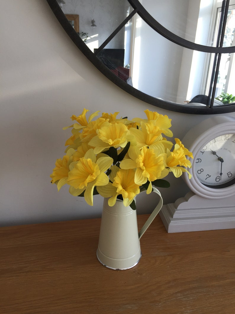 May include: A bouquet of yellow daffodils in a cream-colored metal pitcher on a wooden table.