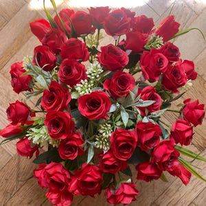 May include: A vibrant bouquet of red roses, accented with green leaves and small white flowers. The arrangement is densely packed, creating a full and lush appearance. The roses are in various stages of bloom, adding depth and texture to the floral display.