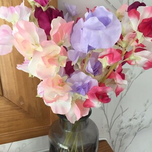 Puede incluir: Un ramo de flores de guisante de olor rosa, morado y rojo en un jarr&oacute;n de vidrio gris oscuro.