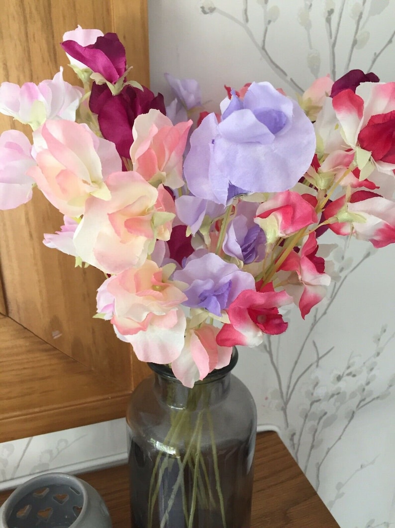 Puede incluir: Un ramo de flores de guisante de olor rosa, morado y rojo en un jarr&oacute;n de vidrio gris oscuro.