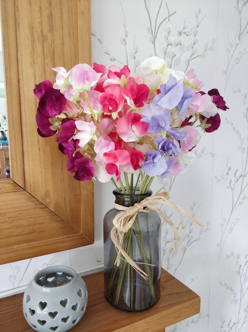 Puede incluir: Un ramo de flores de guisante dulce de color morado, rosa y blanco en un jarr&oacute;n de vidrio transparente con una cinta marr&oacute;n alrededor del cuello.