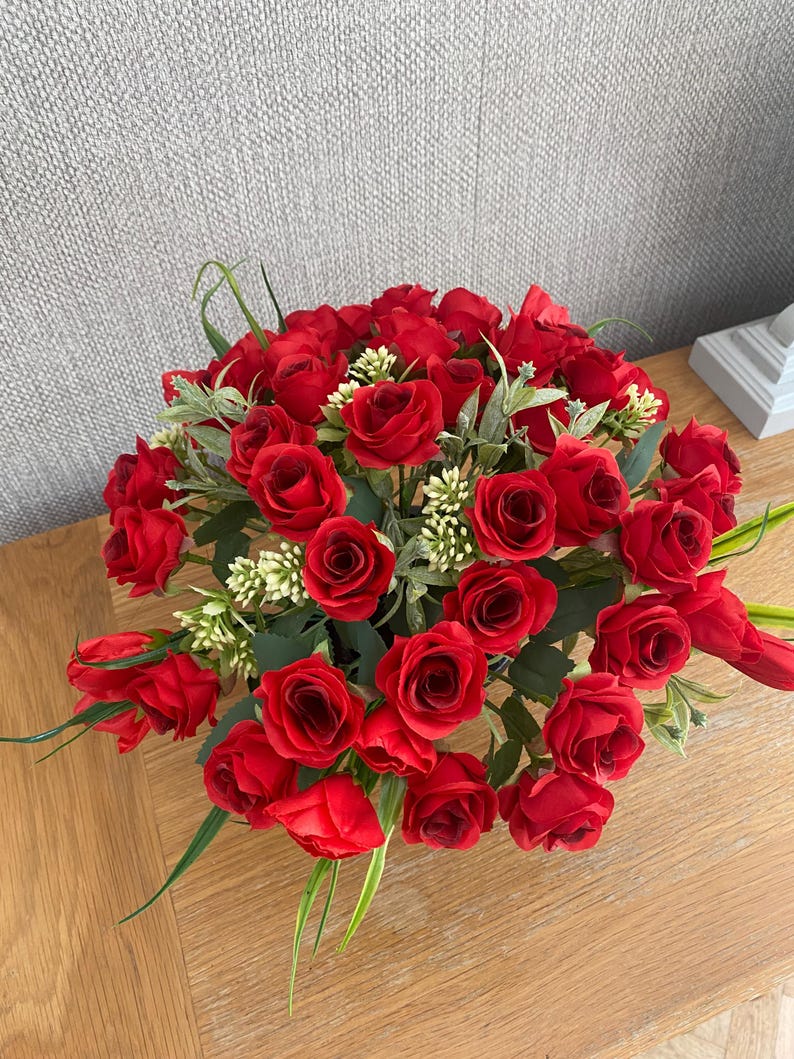 May include: A vibrant arrangement of red roses, accented with green leaves and small white flowers. The bouquet is densely packed, creating a full, lush appearance. The roses are in full bloom, showcasing their rich red color.