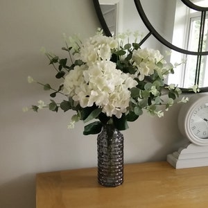 May include: A bouquet of white hydrangeas and green eucalyptus leaves in a grey textured glass vase.
