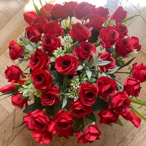 May include: A vibrant bouquet of red roses, accented with green foliage and small white flowers. The arrangement features numerous fully bloomed roses, creating a lush and full display. The roses are a deep, rich red color.