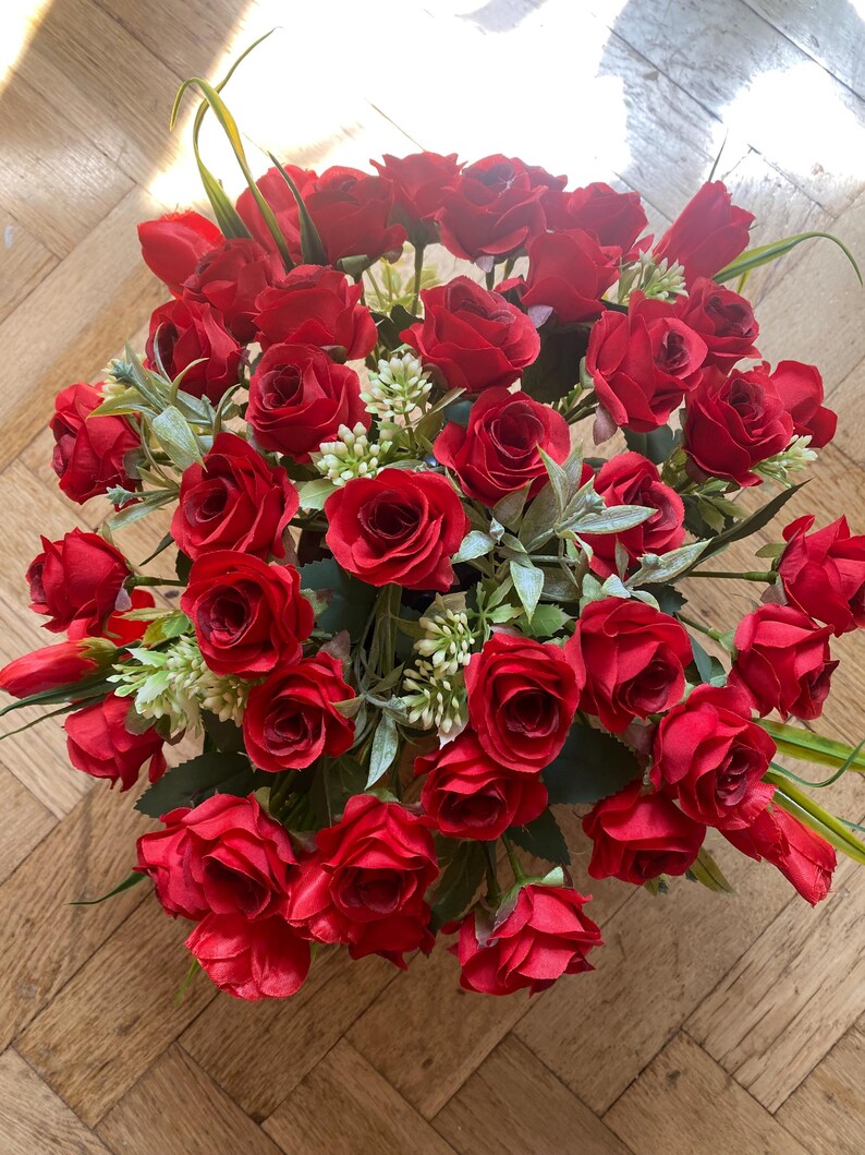 May include: A vibrant bouquet of red roses, accented with green foliage and small white flowers. The arrangement features numerous fully bloomed roses, creating a lush and full display. The roses are a deep, rich red color.