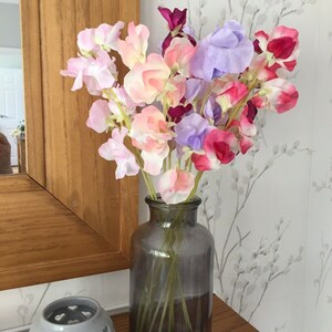 Puede incluir: Un ramo de flores de guisante dulce de color rosa, morado y rojo en un jarr&oacute;n de vidrio transparente con un tinte gris oscuro. El jarr&oacute;n est&aacute; sobre una superficie de madera.