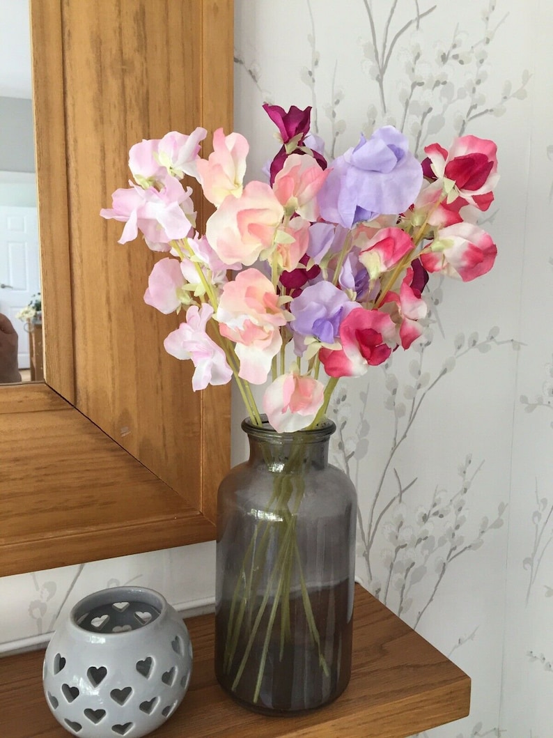 Puede incluir: Un ramo de flores de guisante dulce de color rosa, morado y rojo en un jarr&oacute;n de vidrio transparente con un tinte gris oscuro. El jarr&oacute;n est&aacute; sobre una superficie de madera.