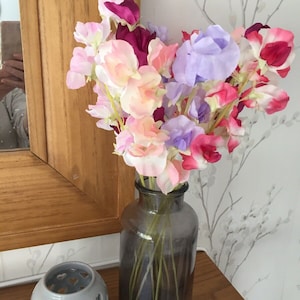 Puede incluir: Un ramo de flores de guisante de olor rosa, p&uacute;rpura y rojo en un jarr&oacute;n de vidrio gris oscuro. El jarr&oacute;n est&aacute; sobre una repisa de madera.