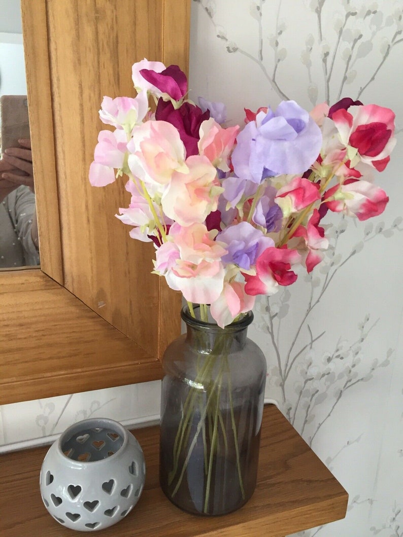 Puede incluir: Un ramo de flores de guisante de olor rosa, p&uacute;rpura y rojo en un jarr&oacute;n de vidrio gris oscuro. El jarr&oacute;n est&aacute; sobre una repisa de madera.