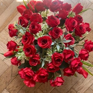 May include: A vibrant bouquet of red roses, interspersed with green foliage and small white flowers. The arrangement is densely packed, creating a full and lush appearance. The roses are in full bloom, showcasing their rich red color.