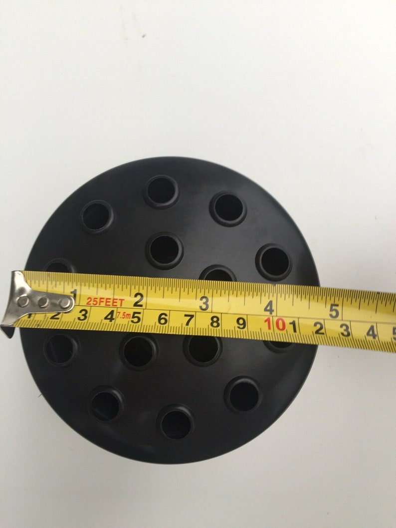 May include: A black floral foam holder with multiple holes for arranging flowers. A yellow tape measure shows the holder is approximately 4 inches in diameter. The holder is on a white surface.