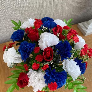 May include: A colourful floral arrangement with red roses, white carnations, and blue carnations. Small red flowers and green leaves are also present. The flowers are arranged in a bouquet.