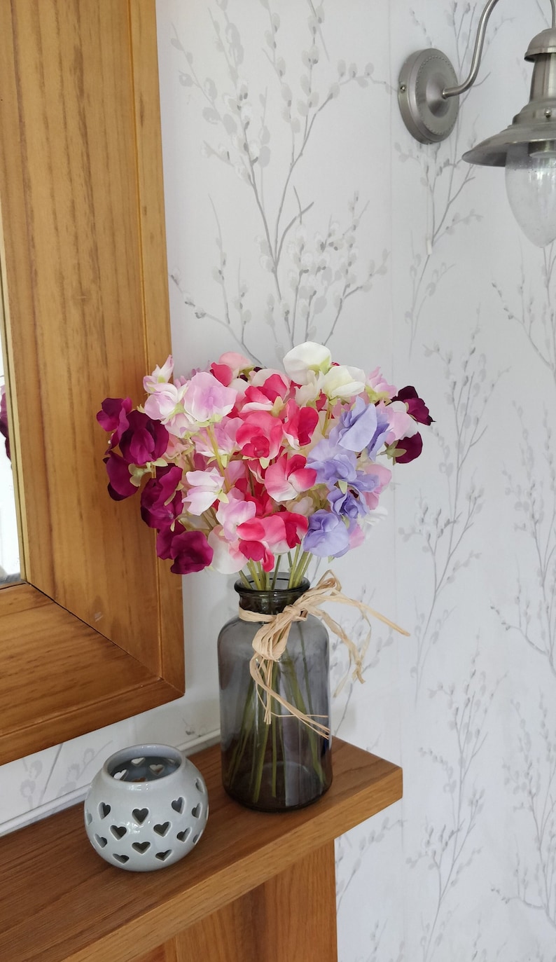 Puede incluir: Un ramo de flores de guisante de olor de color morado, rosa y blanco en un jarr&oacute;n de vidrio transparente con un lazo de cuerda marr&oacute;n. El jarr&oacute;n est&aacute; sobre una repisa de madera.