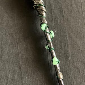 Peut inclure: Une baguette artisanale avec une pointe en cristal clair et un visage sculpté sur le manche. La baguette gris foncé est enveloppée de fil vert et ornée de perles vertes en forme de feuille de verre. Un objet mystique unique.