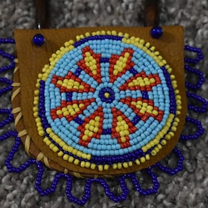 May include: A beaded Native American pouch with a blue, yellow, and red geometric design. The pouch is made of leather and has a fringe of blue beads.