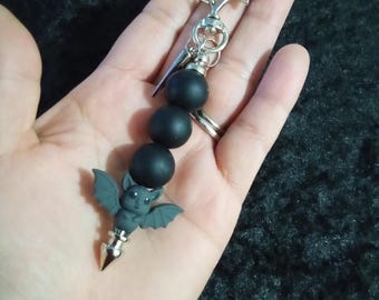 May include: A silver-toned bat-shaped keychain with a black beaded accent and a small gray bat charm. The charm has a silver spike at the bottom. The keychain is held in a hand.