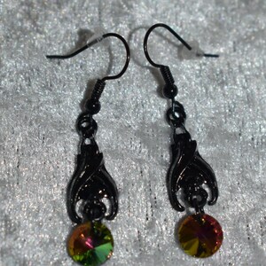 May include: A pair of black metal drop earrings featuring a bat-shaped charm suspended from a hook wire. Each earring is accented with a round, faceted crystal that displays iridescent green, yellow, and reddish-pink hues at the base.