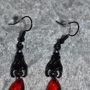 May include: A pair of black metal earrings featuring a hanging bat design with a dangling red teardrop-shaped bead. The earrings are attached to black metal fishhook wires with small clear plastic backings for secure placement during wear.